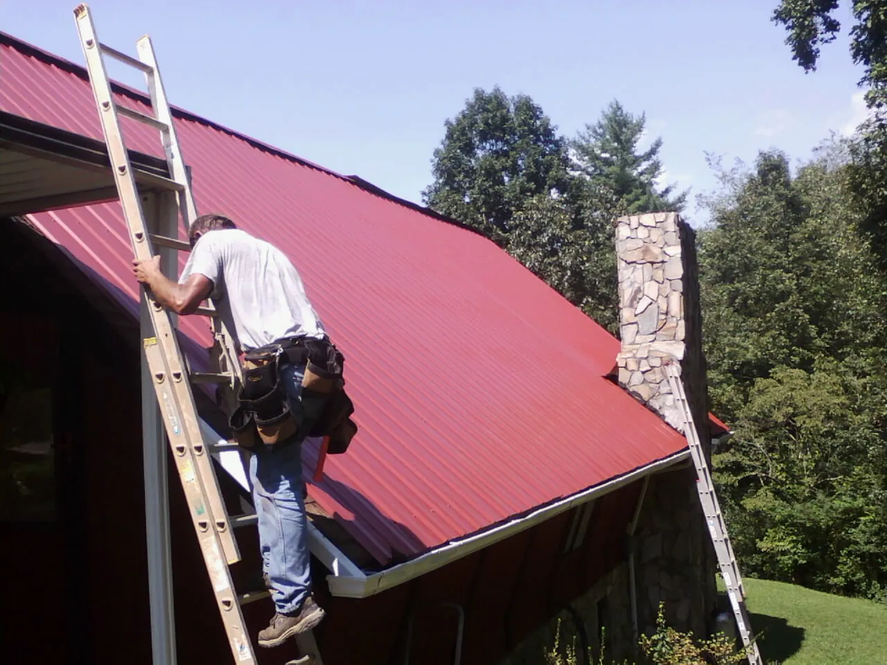Roofer on a ladder beside a red metal roof and stone chimney — Soffit & Fascia Repair by Roofing Brothers in Coosawhatchie